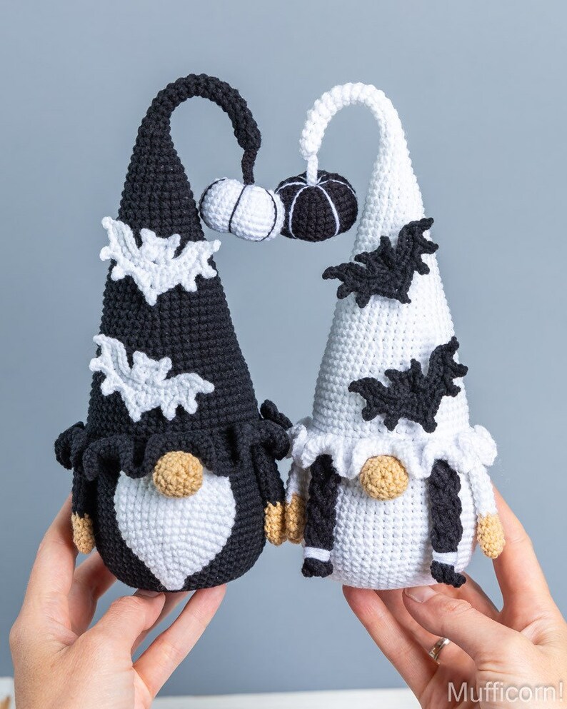 May include: Two crocheted gnomes dressed in black and white with bat and pumpkin accents. The gnomes are holding a small crocheted pumpkin.