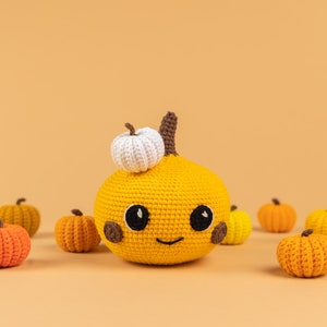 May include: A crocheted pumpkin with a smiling face and a small white crocheted pumpkin on top. The pumpkin is surrounded by smaller crocheted pumpkins in various shades of orange and yellow.