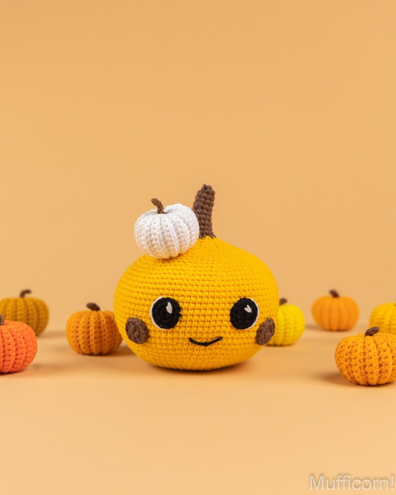 May include: A crocheted pumpkin with a smiling face and a small white crocheted pumpkin on top. The pumpkin is surrounded by smaller crocheted pumpkins in various shades of orange and yellow.