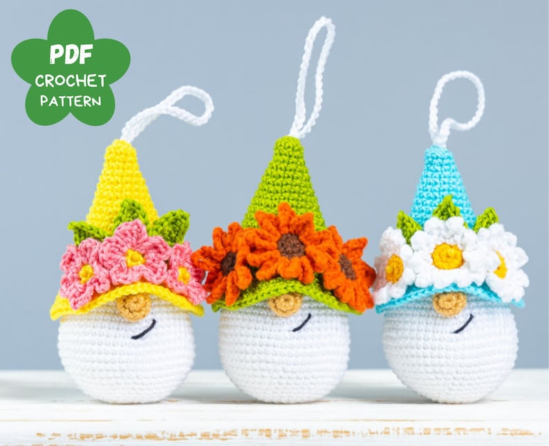 May include: Three crocheted gnomes with different colored hats and flower crowns. The gnome on the left has a yellow hat with pink flowers, the middle gnome has a green hat with orange flowers, and the gnome on the right has a blue hat with white flowers.