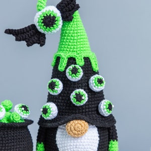 May include: A green and black crocheted gnome with a pointy hat decorated with googly eyes. The gnome has a white beard and a brown nose. The gnome is wearing a black hat with a green brim and a black bat wing on the side.