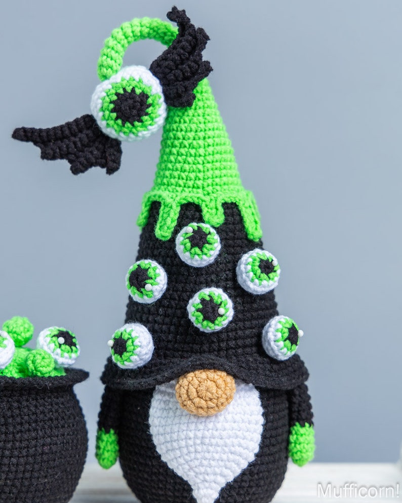 May include: A green and black crocheted gnome with a pointy hat decorated with googly eyes. The gnome has a white beard and a brown nose. The gnome is wearing a black hat with a green brim and a black bat wing on the side.