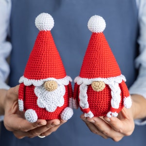 May include: Two crocheted gnomes in red and white, with white pom-poms on their hats. The gnomes are holding hands and have white beards and braids.
