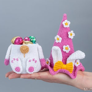 May include: Two crocheted Easter baskets, one shaped like a bunny and the other like a hat with bunny ears. The bunny basket is white with pink accents and is filled with chocolate eggs. The hat basket is pink with white flowers and a yellow bow.