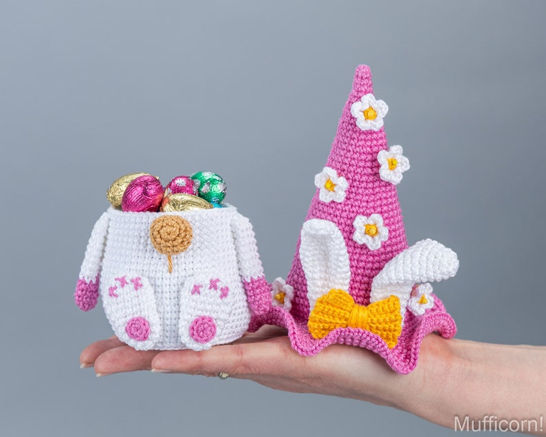 May include: Two crocheted Easter baskets, one shaped like a bunny and the other like a hat with bunny ears. The bunny basket is white with pink accents and is filled with chocolate eggs. The hat basket is pink with white flowers and a yellow bow.