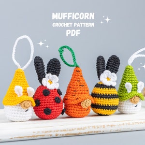 Könnte beinhalten: Fünf gehäkelte Amigurumi-Anhänger in Form von Gnomen mit verschiedenen farbigen Hüten. Die Gnomen tragen Hüte, die gelb, rot, orange, schwarz-gelb gestreift und grün sind. Die Gnomen sitzen auf einer weißen Holzoberfläche. Der Text "MUFFICORN CROCHET PATTERN PDF" befindet sich über den Gnomen.