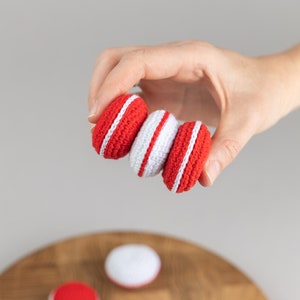 Puede incluir: Tres macarons de ganchillo hechos a mano en rojo y blanco son sostenidos por una mano. Los macarons tienen un exterior rojo con una raya blanca alrededor del centro. Un macaron es blanco con una raya roja. El fondo es gris claro.