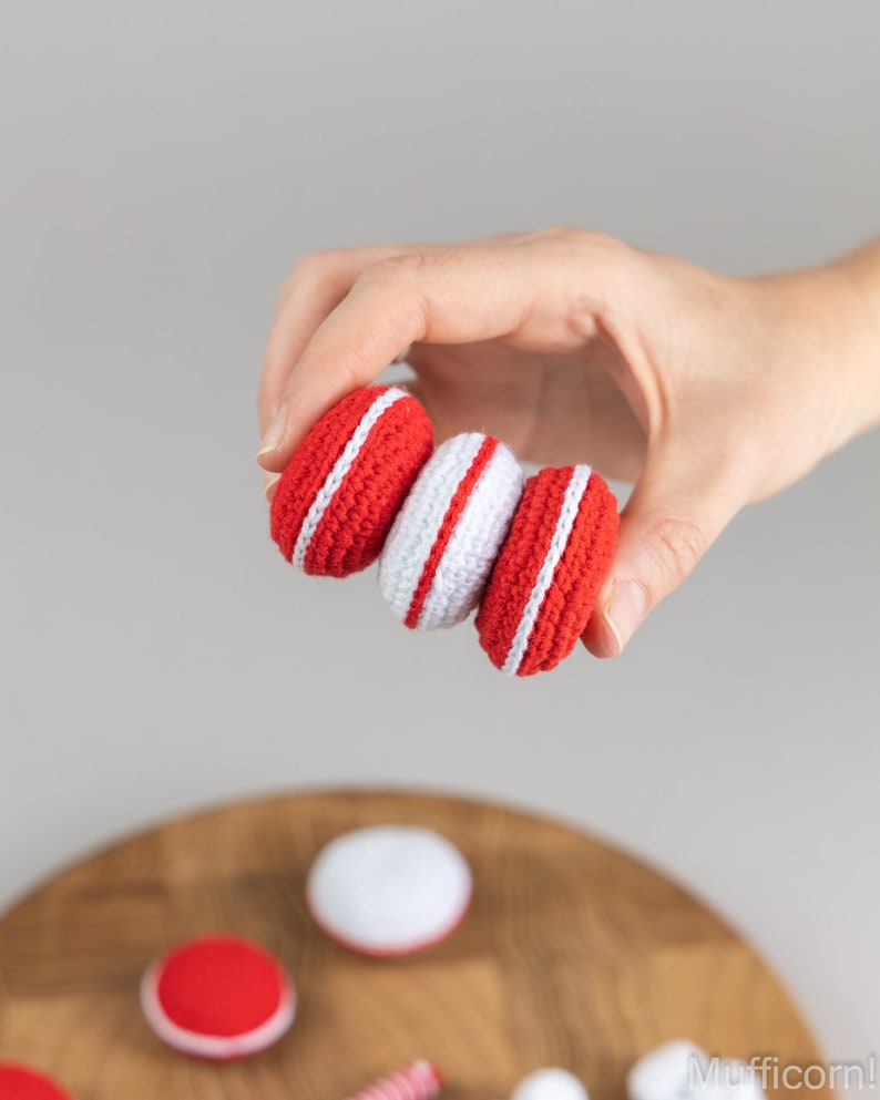 Puede incluir: Tres macarons de ganchillo hechos a mano en rojo y blanco son sostenidos por una mano. Los macarons tienen un exterior rojo con una raya blanca alrededor del centro. Un macaron es blanco con una raya roja. El fondo es gris claro.