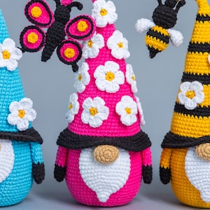 May include: Three crocheted gnomes with different colored hats and decorations. The gnome on the left has a blue hat with white flowers, the middle gnome has a pink hat with white flowers and a black butterfly, and the gnome on the right has a yellow hat with black stripes and a bee.