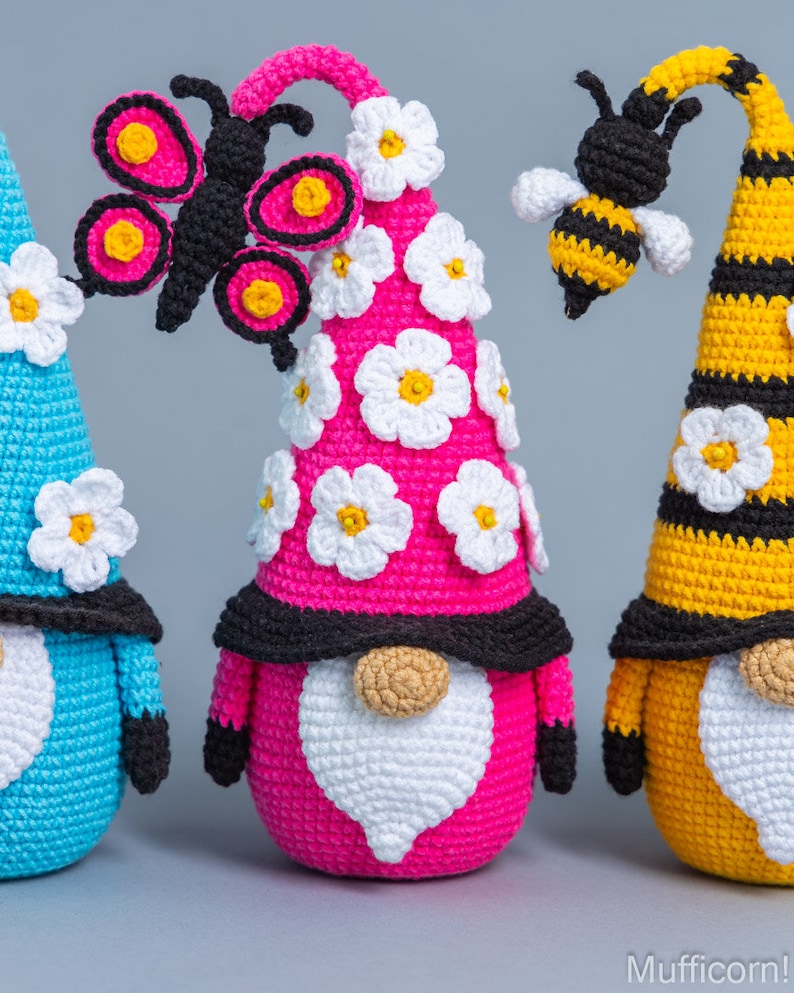 May include: Three crocheted gnomes with different colored hats and decorations. The gnome on the left has a blue hat with white flowers, the middle gnome has a pink hat with white flowers and a black butterfly, and the gnome on the right has a yellow hat with black stripes and a bee.