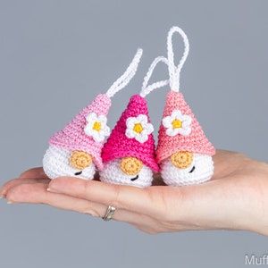 May include: Three handmade crochet gnomes with white bodies, pink hats, and small white daisy accents. Each gnome has a small, tan-colored nose and a white string for hanging. The gnomes are held in a hand against a gray background.
