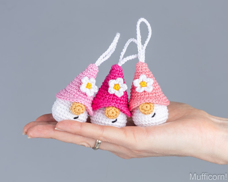 May include: Three handmade crochet gnomes with white bodies, pink hats, and small white daisy accents. Each gnome has a small, tan-colored nose and a white string for hanging. The gnomes are held in a hand against a gray background.