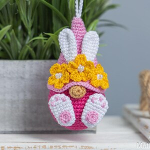 May include: A pink crocheted gnome with white bunny ears and yellow flower accents. The gnome is wearing a pink hat with white bunny ears and a yellow flower crown. The gnome is sitting on a white surface.