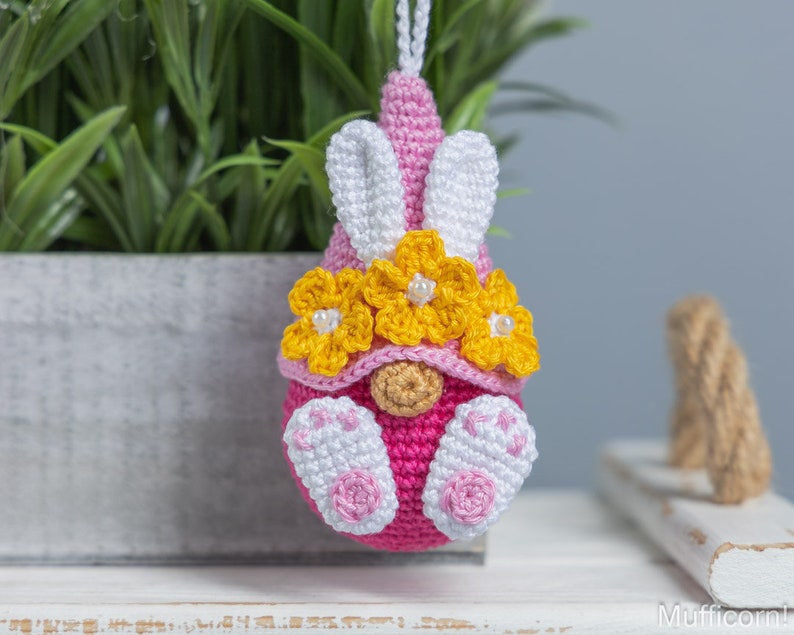 May include: A pink crocheted gnome with white bunny ears and yellow flower accents. The gnome is wearing a pink hat with white bunny ears and a yellow flower crown. The gnome is sitting on a white surface.