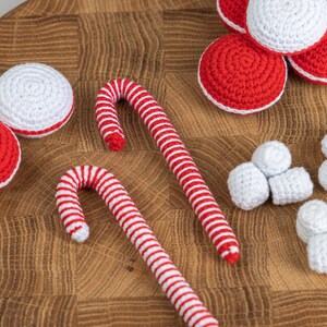 Puede incluir: Una colecci&oacute;n de adornos navide&ntilde;os hechos a mano con ganchillo, que incluyen bastones de caramelo a rayas rojas y blancas, adornos redondos rojos y blancos y peque&ntilde;os trozos blancos en forma de malvavisco. Los art&iacute;culos est&aacute;n dispuestos sobre una superficie de madera.