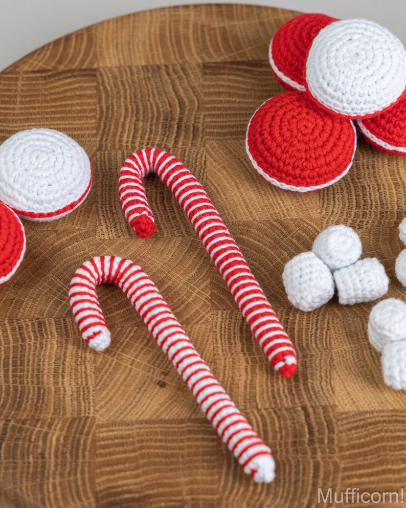 Puede incluir: Una colecci&oacute;n de adornos navide&ntilde;os hechos a mano con ganchillo, que incluyen bastones de caramelo a rayas rojas y blancas, adornos redondos rojos y blancos y peque&ntilde;os trozos blancos en forma de malvavisco. Los art&iacute;culos est&aacute;n dispuestos sobre una superficie de madera.