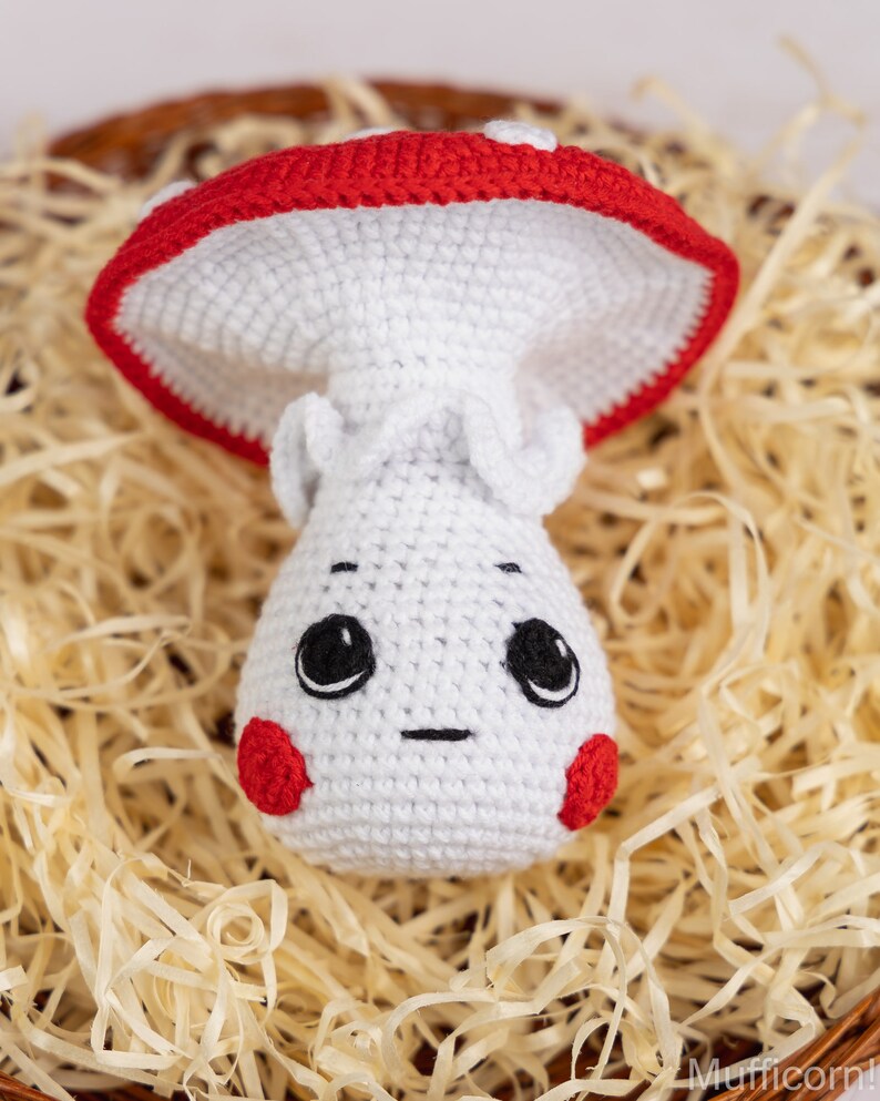 May include: A crocheted mushroom toy with a white body, red cap, and a cute embroidered face. The mushroom is sitting in a basket of shredded paper.