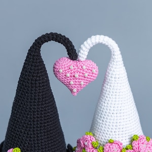 May include: Two crocheted cone-shaped figures with a pink heart in the center. The figure on the left is black and has a pink rose at the base. The figure on the right is white and has a row of pink roses at the base.