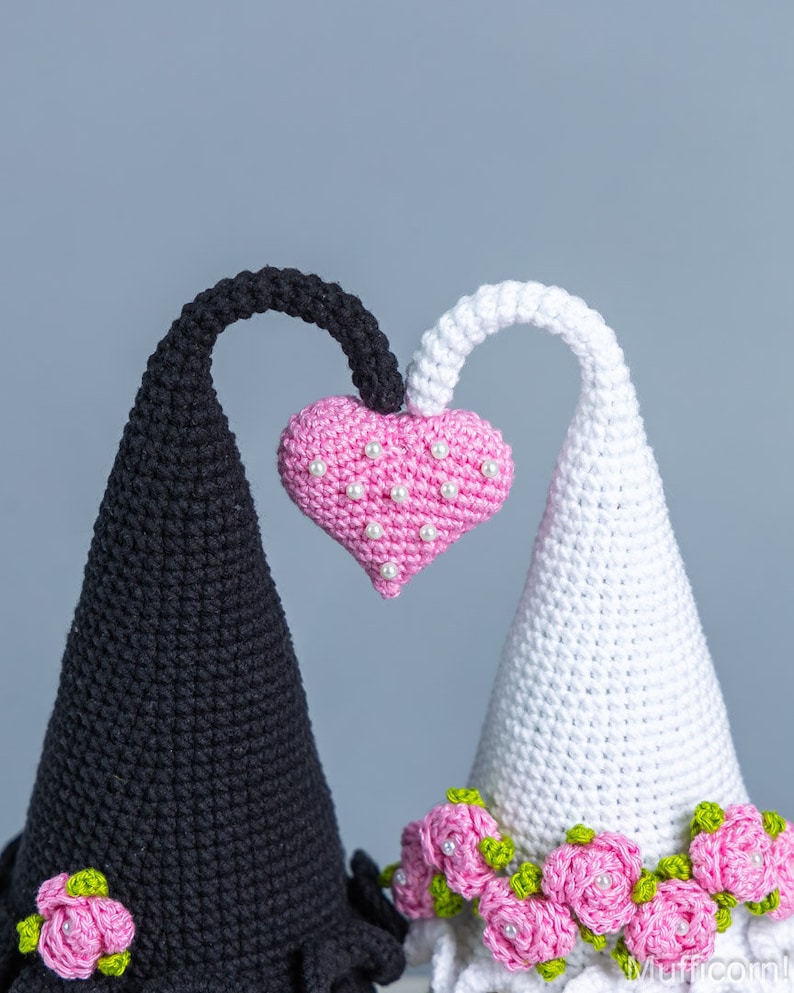 May include: Two crocheted cone-shaped figures with a pink heart in the center. The figure on the left is black and has a pink rose at the base. The figure on the right is white and has a row of pink roses at the base.