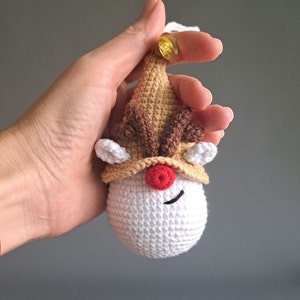 May include: A crocheted reindeer ornament with a white face, red nose, and brown antlers. The ornament is brown and white and has a gold bell on top.