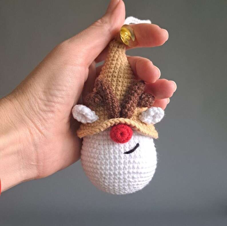 May include: A crocheted reindeer ornament with a white face, red nose, and brown antlers. The ornament is brown and white and has a gold bell on top.