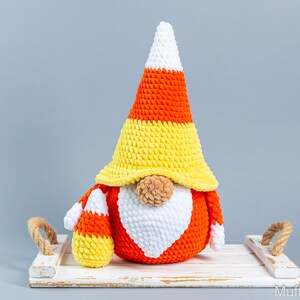 May include: A crocheted candy corn gnome with a white beard and a yellow, orange, and white hat. The gnome is sitting on a white wooden tray with rope handles.