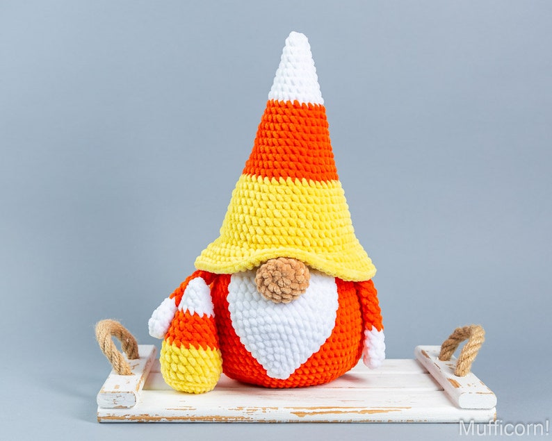 May include: A crocheted candy corn gnome with a white beard and a yellow, orange, and white hat. The gnome is sitting on a white wooden tray with rope handles.