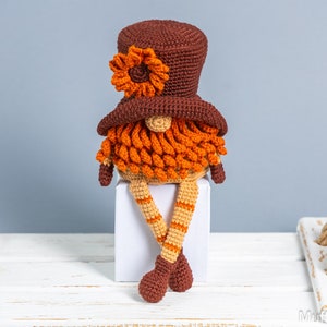May include: A crocheted gnome with a brown hat and orange beard sits on a white block. The gnome has orange and brown striped legs.