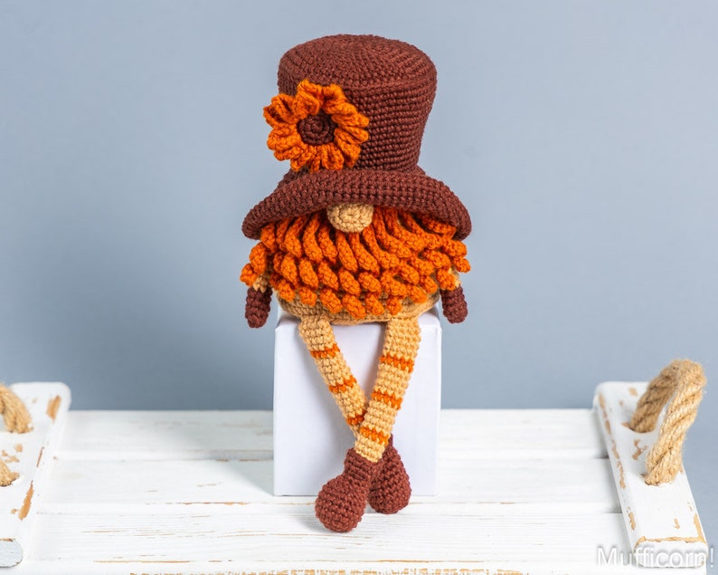 May include: A crocheted gnome with a brown hat and orange beard sits on a white block. The gnome has orange and brown striped legs.