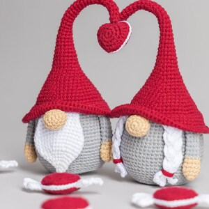 May include: Two crocheted gnomes with red hats and gray bodies. The gnomes are standing with their heads tilted towards each other, forming a heart shape with their hats. There are small red and white candy-like objects scattered around the gnomes.
