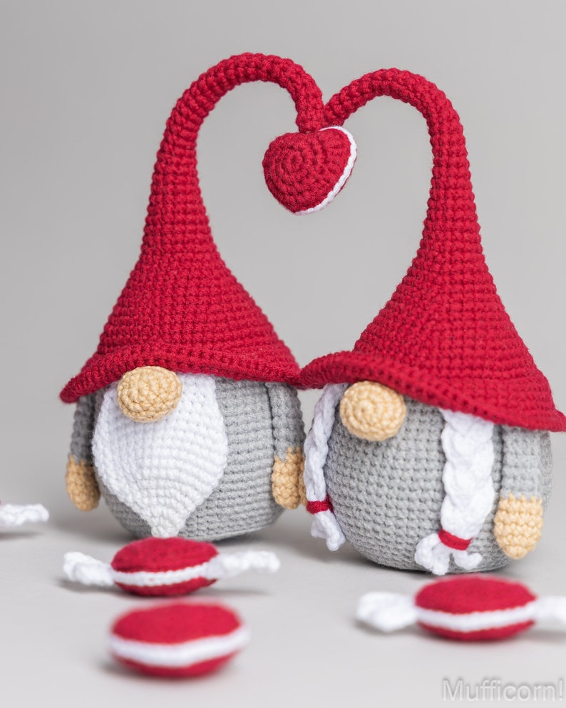 May include: Two crocheted gnomes with red hats and gray bodies. The gnomes are standing with their heads tilted towards each other, forming a heart shape with their hats. There are small red and white candy-like objects scattered around the gnomes.