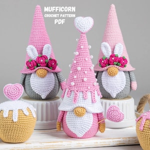 Può includere: Tre gnomi all'uncinetto rosa con barbe bianche e orecchie di coniglio. Gli gnomi indossano cappelli rosa e bianchi con fiori e perle. Gli gnomi sono seduti su basi bianche. Ci sono due cupcake all'uncinetto con cuori rosa in cima.