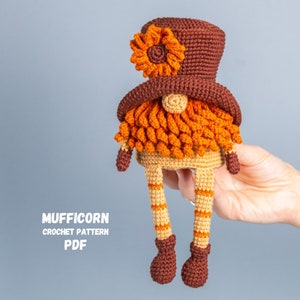 May include: Crocheted gnome with a brown hat and orange beard. The hat has a yellow flower on top. The gnome is wearing brown shoes and has orange and white striped legs. The text "MUFFICORN CROCHET PATTERN PDF" is visible in the image.