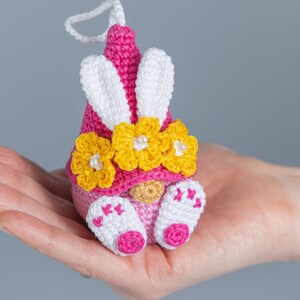 May include: A pink and white crocheted gnome with yellow flower accents and bunny ears. The gnome is holding a small white ball in its hands.