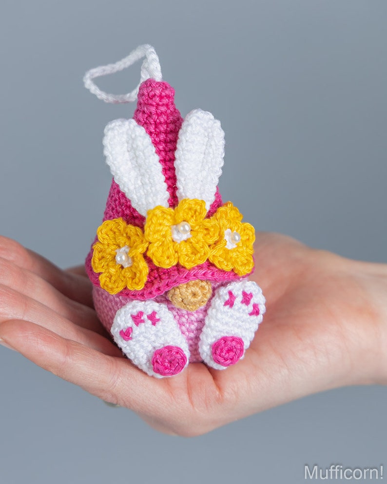 May include: A pink and white crocheted gnome with yellow flower accents and bunny ears. The gnome is holding a small white ball in its hands.