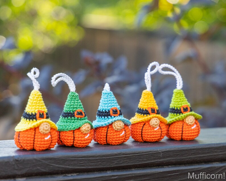 May include: Five crocheted pumpkin gnomes with different colored hats. The gnomes are orange with black eyes and noses. The hats are yellow, green, blue, yellow, and green.