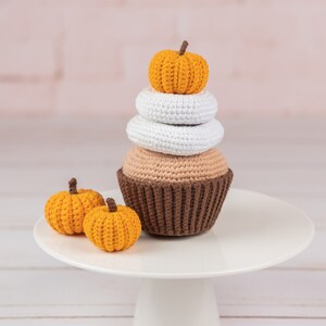 May include: A crocheted cupcake with brown, white, and orange frosting topped with a small orange pumpkin. Two smaller orange pumpkins are on a white cake stand.