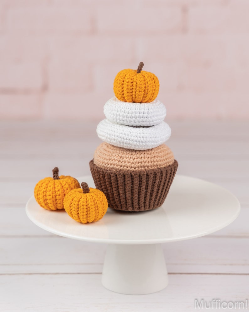 May include: A crocheted cupcake with brown, white, and orange frosting topped with a small orange pumpkin. Two smaller orange pumpkins are on a white cake stand.