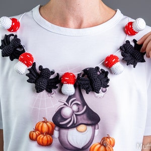 May include: A white t-shirt with a Halloween-themed design featuring a gnome with a spider on its head, pumpkins, and a crocheted garland of bats and red and white mushrooms.