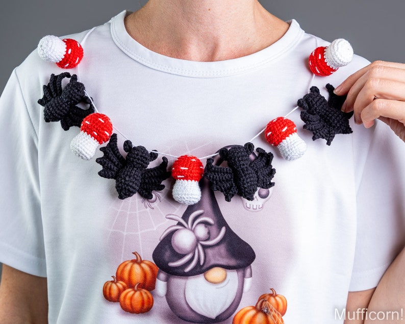 May include: A white t-shirt with a Halloween-themed design featuring a gnome with a spider on its head, pumpkins, and a crocheted garland of bats and red and white mushrooms.