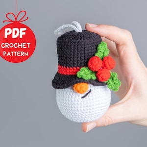 Puede incluir: Un adorno de muñeco de nieve tejido a crochet con un sombrero negro, acebo rojo y verde y una cara blanca. El adorno está siendo sostenido por una mano.