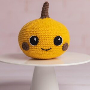 May include: A crocheted yellow pumpkin with a brown stem and a smiling face. The pumpkin has black eyes and brown cheeks. The pumpkin is sitting on a white cake stand.