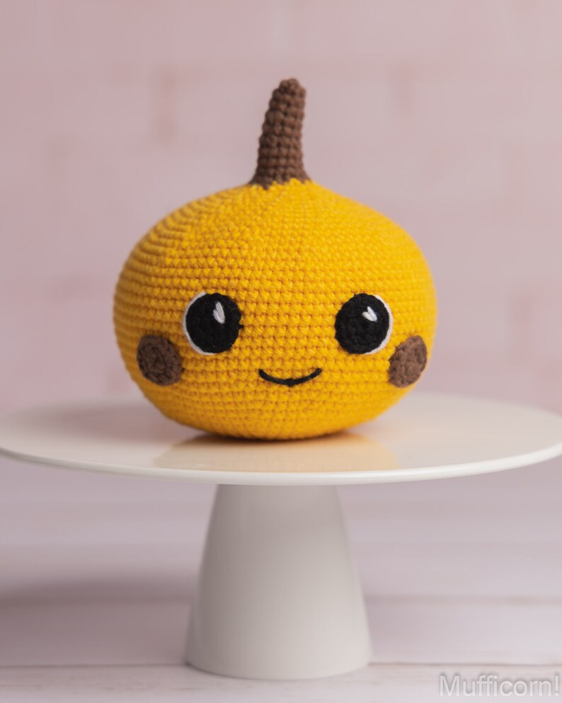 May include: A crocheted yellow pumpkin with a brown stem and a smiling face. The pumpkin has black eyes and brown cheeks. The pumpkin is sitting on a white cake stand.