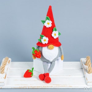 Op de afbeelding: Een gehaakte gnome met een rode muts en witte bloemen, die een rode aardbei vasthoudt. De gnome zit op een wit houten dienblad met een touwhandvat.