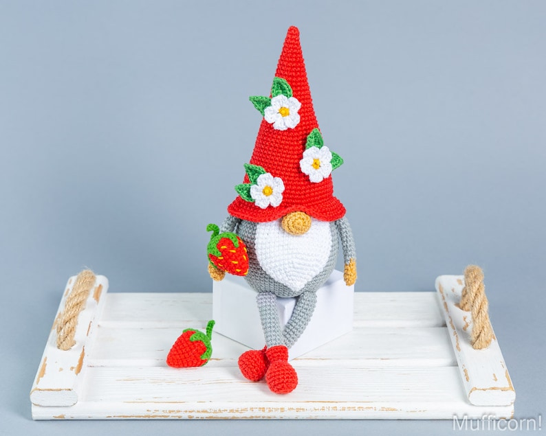 Op de afbeelding: Een gehaakte gnome met een rode muts en witte bloemen, die een rode aardbei vasthoudt. De gnome zit op een wit houten dienblad met een touwhandvat.