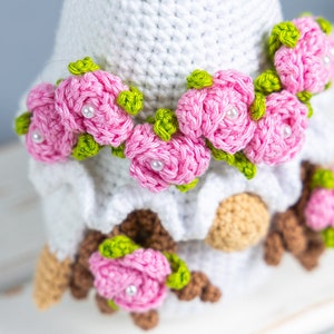 May include: A white crocheted gnome with a pink flower crown and brown crocheted details. The gnome is sitting on a white surface.