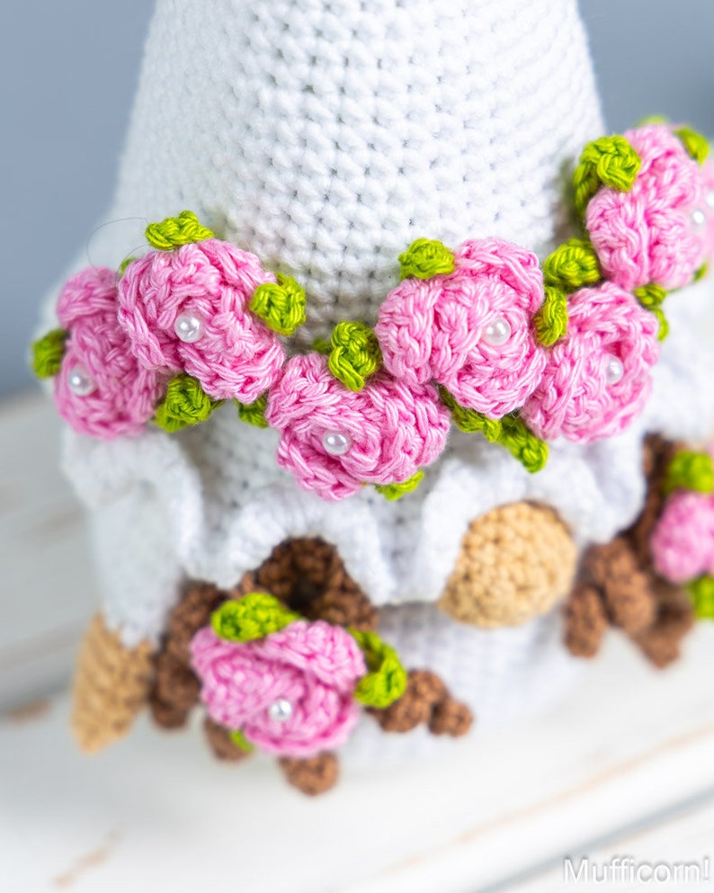 May include: A white crocheted gnome with a pink flower crown and brown crocheted details. The gnome is sitting on a white surface.