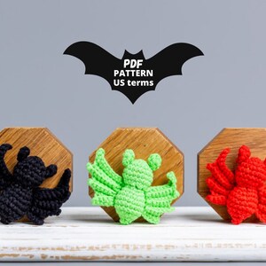 May include: Three crocheted bats in black, green, and red, displayed on wooden stands. The bats are all facing forward with their wings spread out. The text "PDF PATTERN US terms" is displayed above the bats.