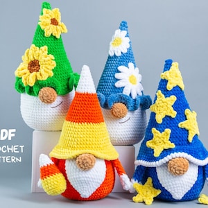 Könnte beinhalten: Vier gehäkelte Gnome mit verschiedenen farbigen Mützen. Die Gnome tragen weiße, grüne, orange und blaue Mützen. Die Mützen sind mit Sonnenblumen, Mais und Sternen verziert. Die Gnome sitzen auf einer weißen Oberfläche. PDF-HÄKELANLEITUNG.