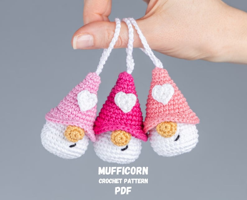 May include: Three handmade crochet gnomes in shades of pink and white, each with a heart-shaped accent on the hat. The gnomes have white bodies, tan noses, and black eyes. The text "MUFFICORN CROCHET PATTERN PDF" is visible.